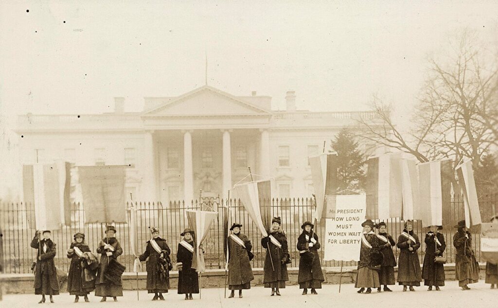 Photograph of fourteen suffragists in overcoats on picket line, holding suffrage banners in front of the White House. One banner reads: "Mr. President How Long Must Women Wait For
